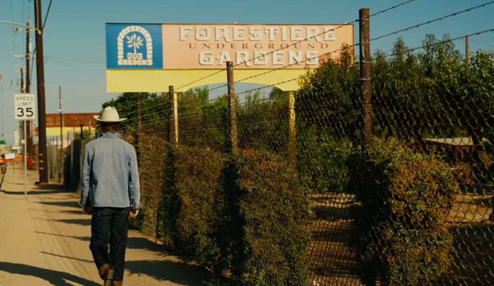 Person in denim and cowboy hat walking by Foretiero Underground Gardens sign and speed limit 35 sign.